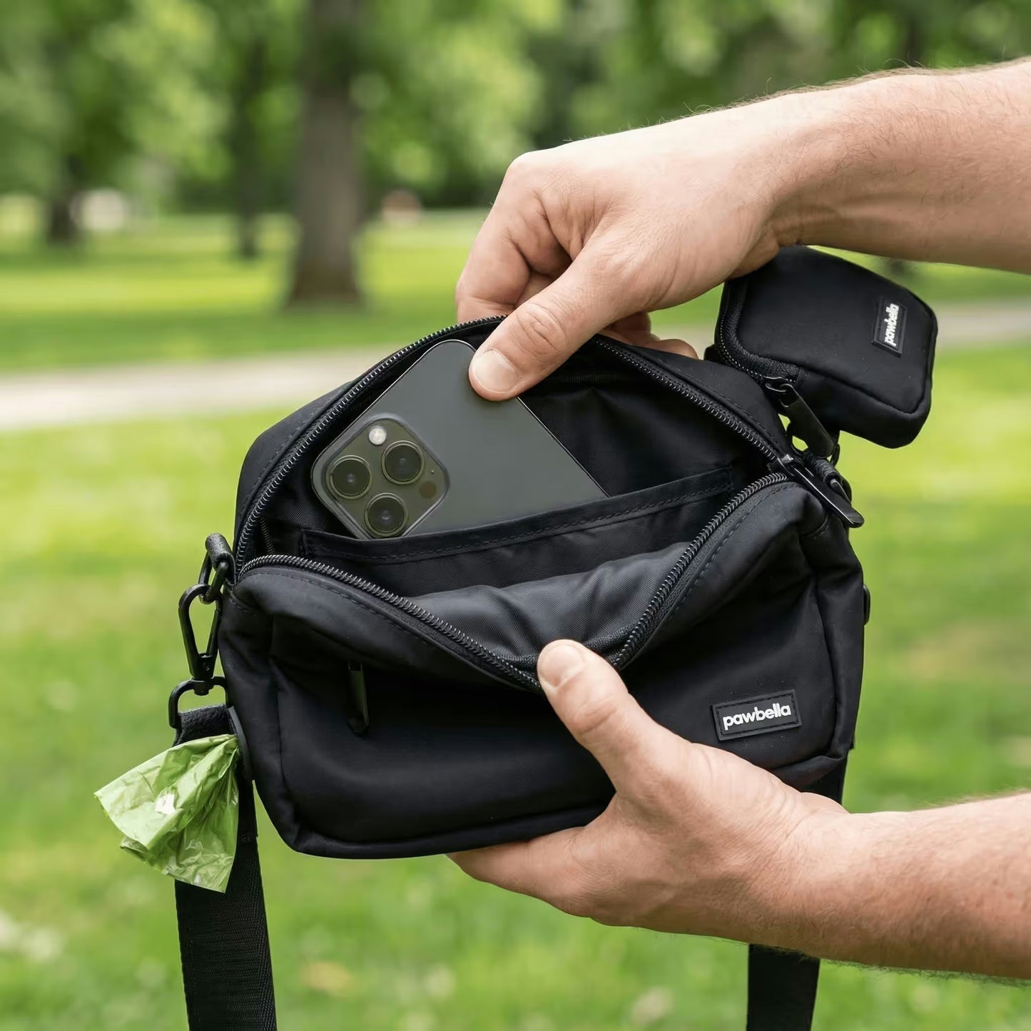 Outdoors in an Australian park with green grass and trees, a person puts a smartphone into the front pocket of a small black Pawbella Crossbody Dog Walking Bag with a pawbell tag.