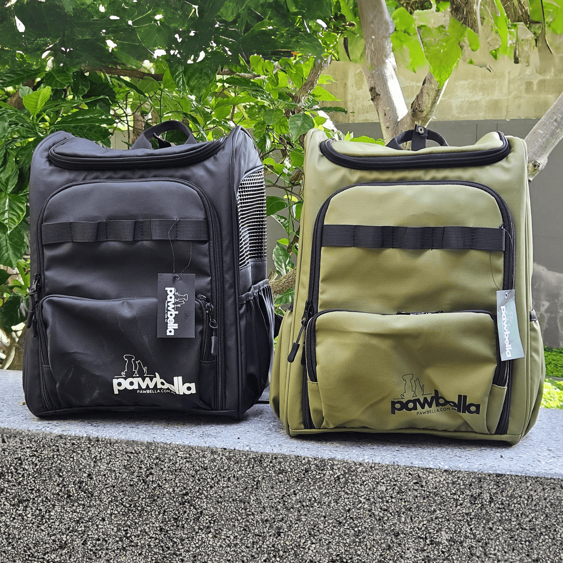 Two Pawbella pet backpacks, one black and one olive, displayed outdoors with lush greenery in the background