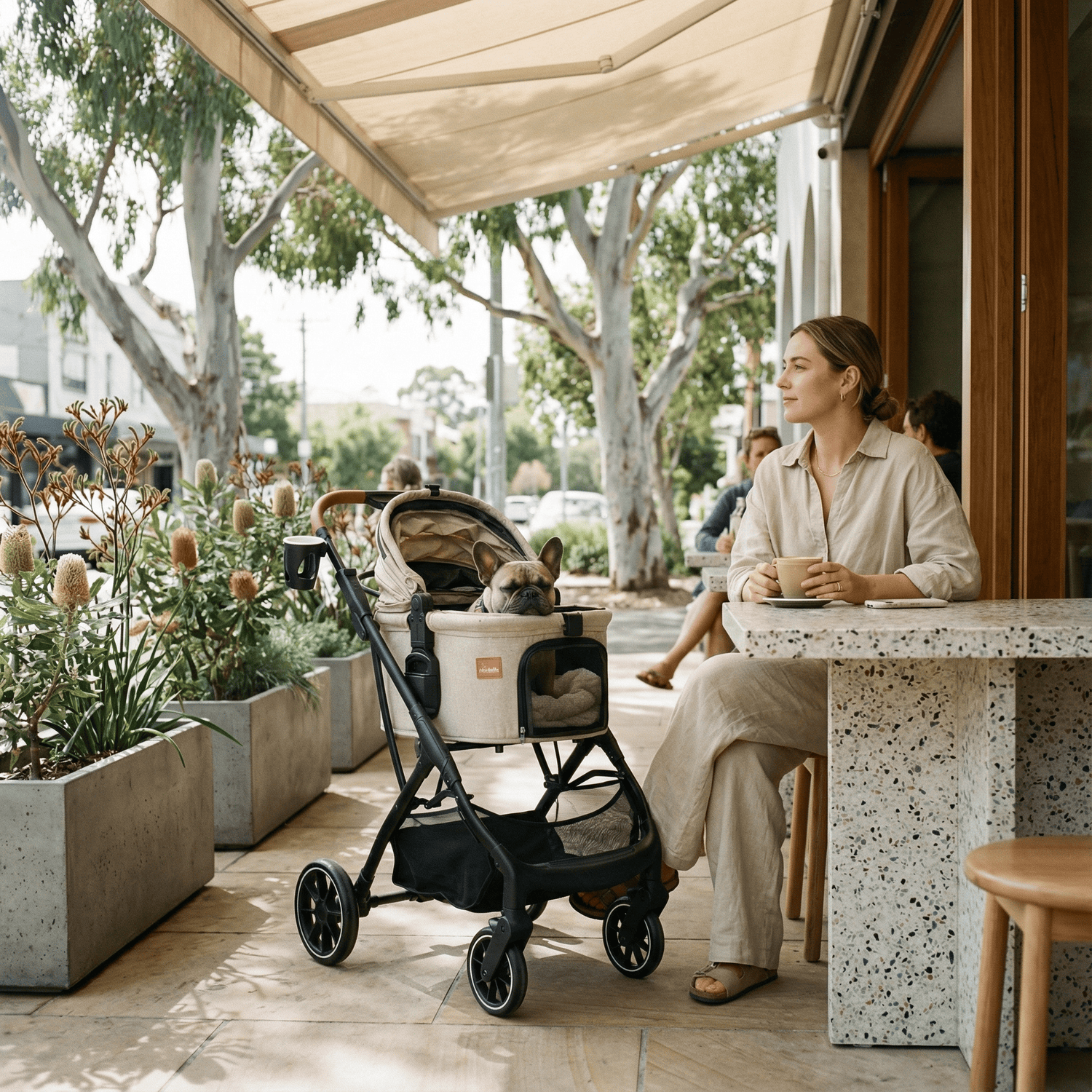 A woman enjoying a coffee break at an outdoor cafe with her French Bulldog resting comfortably in a Pawbella Ultimate Pet Stroller.