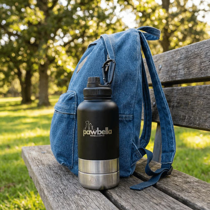A Pawbella 3-in-1 Dog Water Bottle 1L with Food Bowls in black and silver rests on a wooden park bench beside a blue denim pet backpack, with green grass and trees visible on a sunny day in Australia.