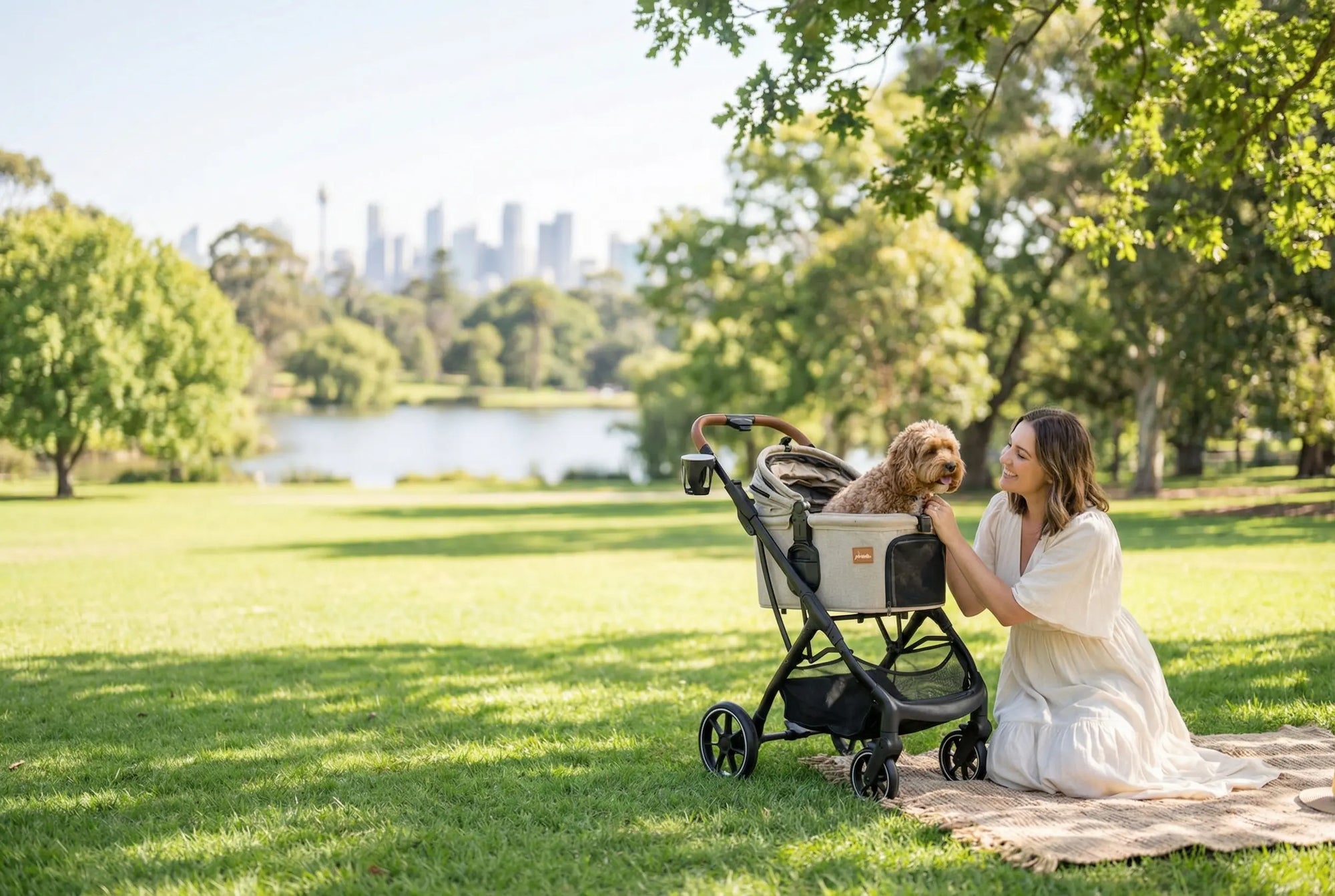 A woman in a white dress kneels on grass in a park, smiling at a small brown dog sitting in a stroller. Trees, a pond, and a distant city skyline are visible in the background under a sunny sky.