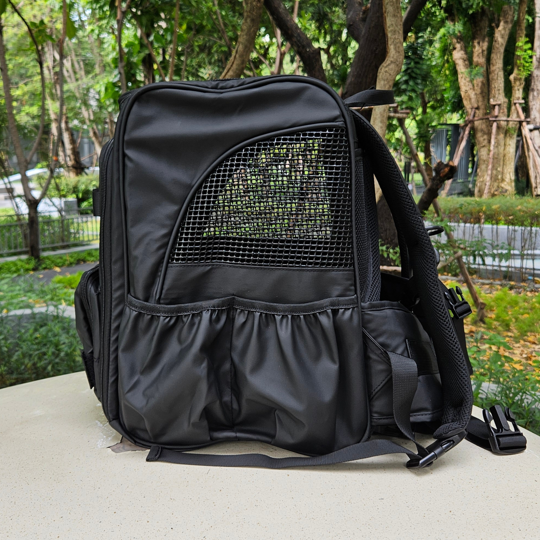 Side view of a black outdoor pet backpack with breathable mesh panel and storage pockets, highlighting durable design and ventilation, set against a green outdoor background.