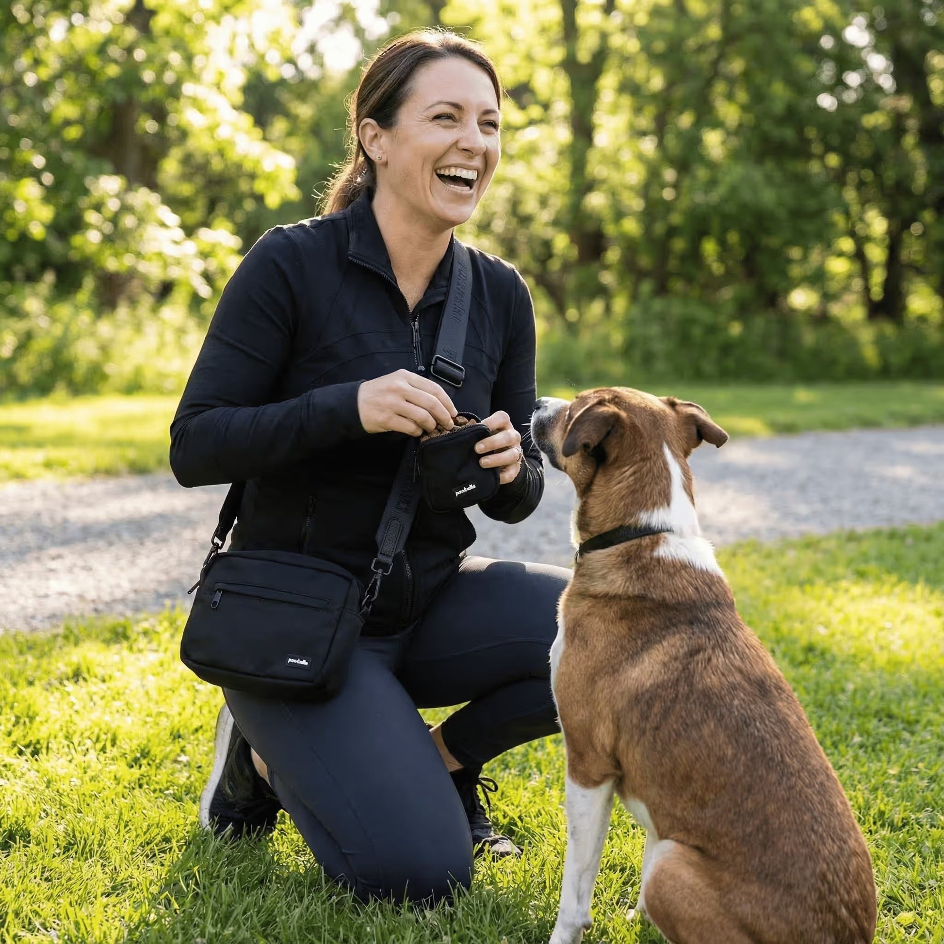 A woman kneels on grass in a sunny Australian park, smiling and training her brown and white dog with treats from the Pawbella Crossbody Dog Walking Bag, while a pet stroller sits nearby.