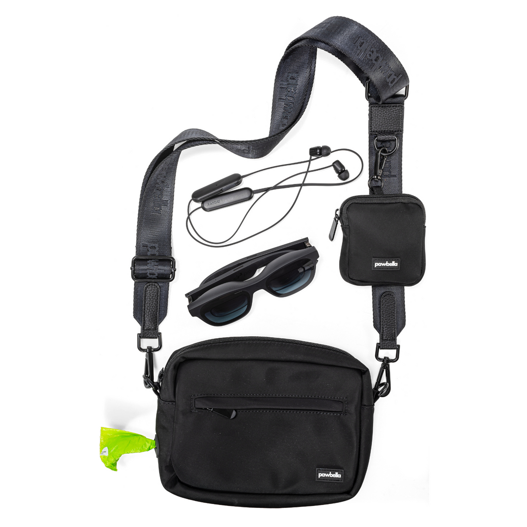 Pawbella dog walking shoulder bag in black with adjustable strap, shown alongside personal accessories including sunglasses, earbuds, and a compact pouch for versatile use