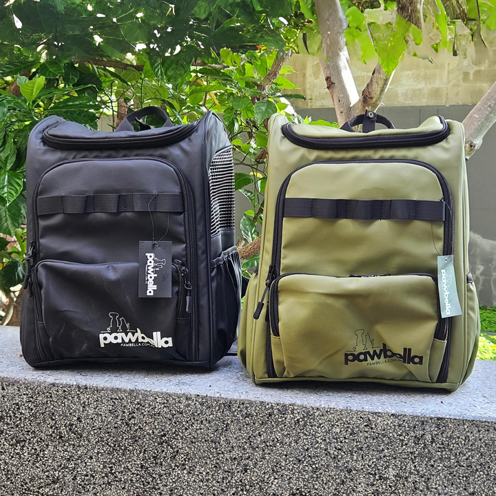 Two Pawbella pet backpacks, one black and one olive, displayed outdoors with lush greenery in the background