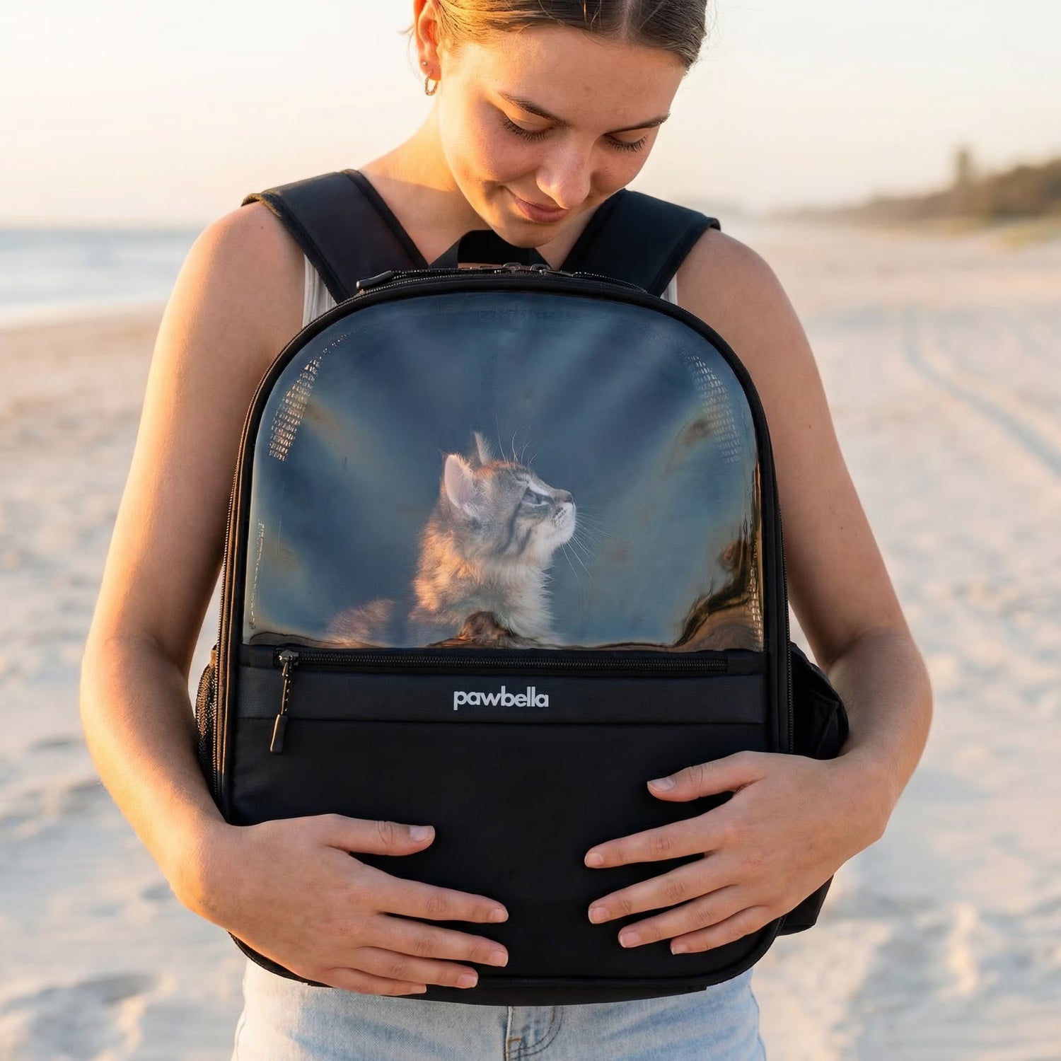 Close-up of a girl on a Gold Coast beach at sunset wearing the Pawbella pet backpack on her front. She looks down lovingly at a kitten inside the ventilated clear window carrier.