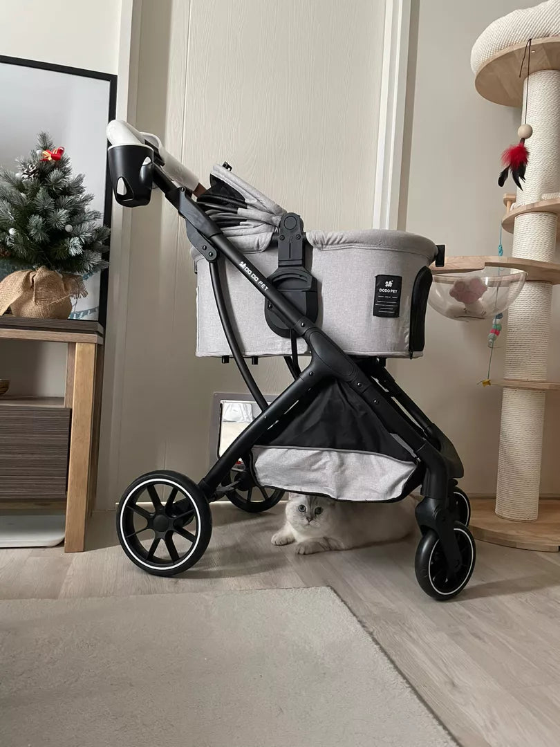 Folded grey pet stroller indoors with a curious cat peeking out from underneath, featuring sleek design elements and positioned near a festive tree